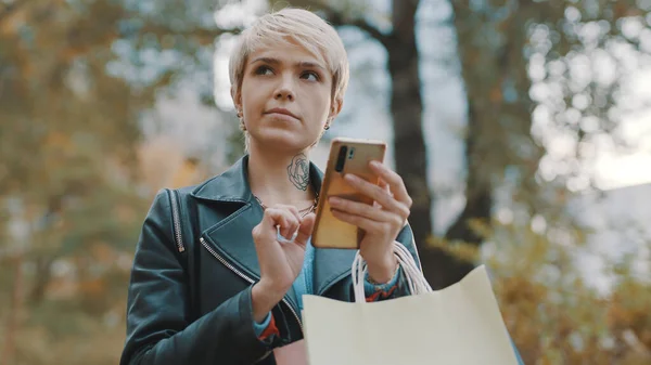 Jovem de cabelos curtos mulher com sacos de compras usando smartphone no parque da cidade — Fotografia de Stock