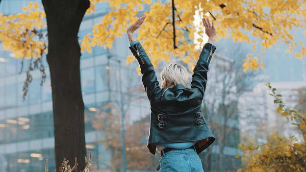 Junge Frau in Lederjacke springt vor Aufregung unter dem herbstlichen Baum im Stadtpark — Stockfoto