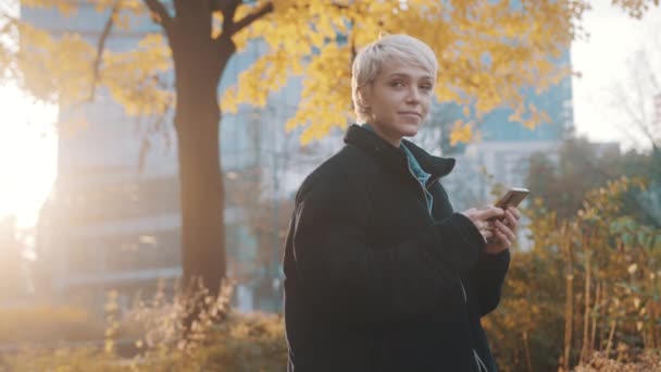 Beyaz tenli, kısa saçlı, sarışın bir kadın. Sonbaharda ofis binasının önündeki parkta telefon kullanıyor. — Stok video