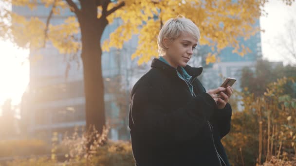 Jonge Kaukasische blonde vrouw met behulp van telefoon in het stadspark in de voorkant van kantoorgebouw in de herfst — Stockvideo