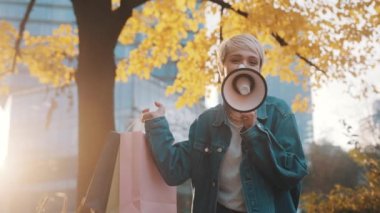 Alışveriş torbalarıyla genç beyaz kadın sonbaharda şehir parkında hoparlörle satış sezonu ilan ediyor.