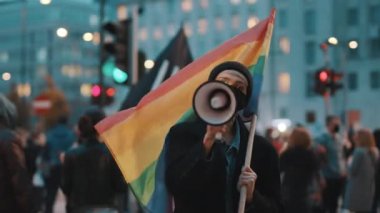Yüz maskeli bir kadın elinde gökkuşağı bayrağı tutarken megafona konuşuyor. Ayrımcılığa karşı gösteri 