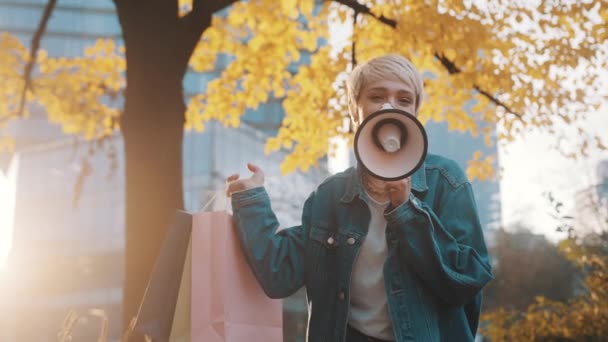 Молода біла жінка з сумками оголошує сезон продажів з гучномовцем у міському парку восени — стокове відео