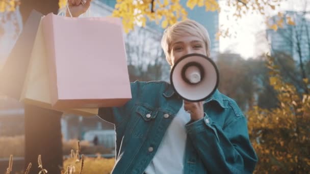 Jonge blonde vrouw met boodschappentassen kondigt verkoopseizoen aan met luidspreker in het stadspark in de herfst — Stockvideo