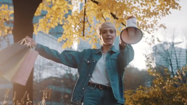November verkoop. Jonge vrouw met boodschappentassen kondigt verkoopseizoen aan met megafoon in het stadspark in de herfst — Stockvideo