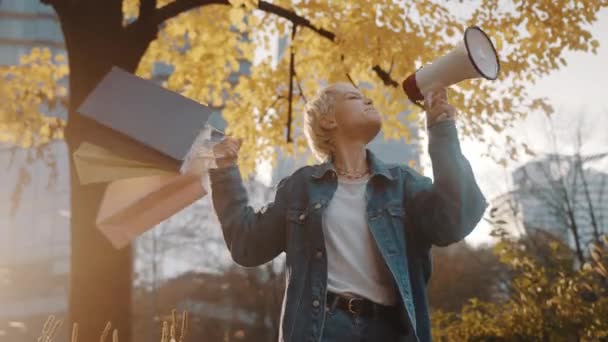 Listopadowa sprzedaż. Blondynka z torbami na zakupy ogłaszająca sezon sprzedaży z megafonem w parku miejskim jesienią — Wideo stockowe