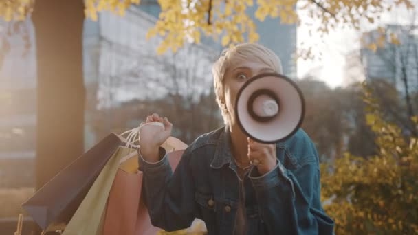 Jonge blonde vrouw met boodschappentassen kondigt verkoopseizoen aan met megafoon in het stadspark in de herfst — Stockvideo