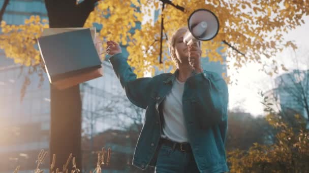 Jonge blonde vrouw met boodschappentassen kondigt verkoopseizoen aan met luidspreker in het stadspark in de herfst — Stockvideo