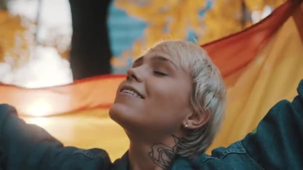 Schöne junge Frau mit kurzen blonden Haaren schwenkt Regenbogenfahne im Park — Stockvideo