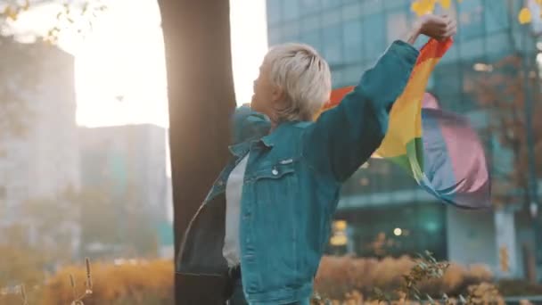 Vacker ung kvinna med kort blont hår spinning och viftande regnbåge flagga i parken — Stockvideo