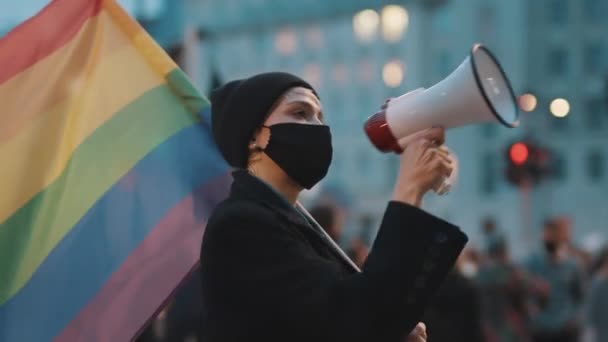 Женщина в маске говорит в мегафон, держа в руках радужный флаг. Демонстрация против дикриминации — стоковое видео