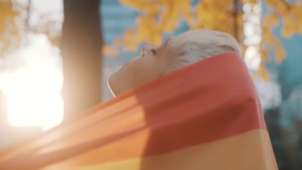 Hübsche junge Frau mit kurzen blonden Haaren, die ihren Kopf mit Regenbogenfahne im Park bedeckt — Stockvideo