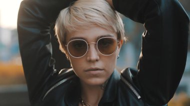 Parlak saçlı, güneş gözlüklü, elleri başının üstünde duran neşeli genç bir kadının portresini yakından çek. İnsanlar ve tarz kavramı.