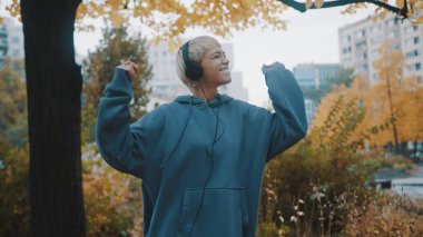 Genç sarışın, kısa saçlı, kapüşonlu kadın sonbaharda şehir parkında dans ve dans müziği dinliyor.