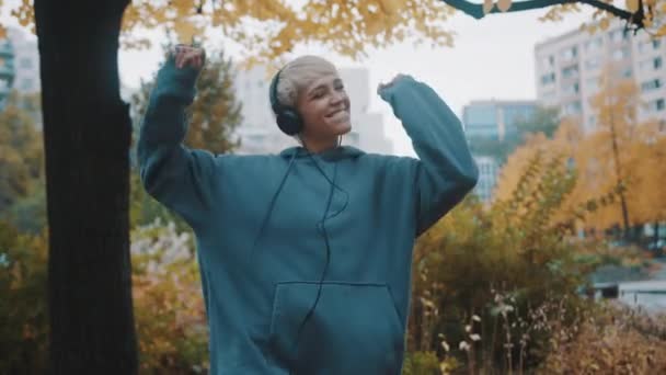 Kapüşonlu mutlu genç sarışın kadın sonbaharda şehir parkında müzik dinliyor ve dans ediyor. — Stok video