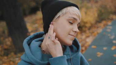 Siyah şapkalı deli, kendine güvenen genç bir kadın parkta kulaklık takarak akıllı telefondan müzik dinliyor.