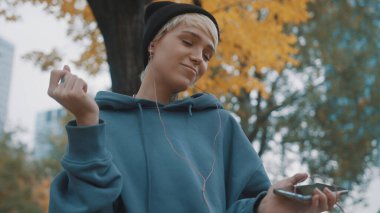 Siyah şapkalı deli, kendine güvenen genç bir kadın parkta kulaklık takarak akıllı telefondan müzik dinliyor.