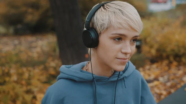 Portret van een jonge blonde vrouw die naar de muziek in het park luistert met een koptelefoon op herfstdag — Stockfoto