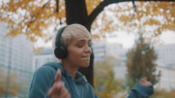 Close up. Happy young woman in the hoodie dancing in the park with headphones in autumn — Stock Video
