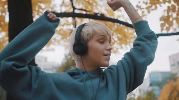 秋の日にヘッドフォンを使用して公園で音楽を聴く若いブロンドの女性の肖像画 — ストック動画