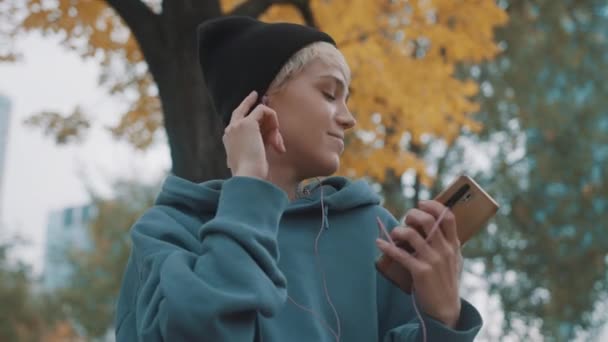 Galen självsäker ung kvinna med vinter svart hatt lyssna på musik från smartphone med hörlurar i parken — Stockvideo