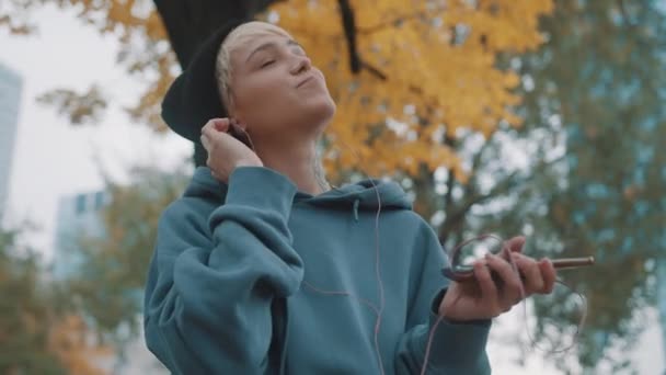 Siyah şapkalı deli, kendine güvenen genç bir kadın parkta kulaklık takarak akıllı telefondan müzik dinliyor. — Stok video