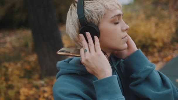 Die junge blonde Frau hört im Park über Kopfhörer Musik von ihrem Smartphone. Porträt — Stockvideo
