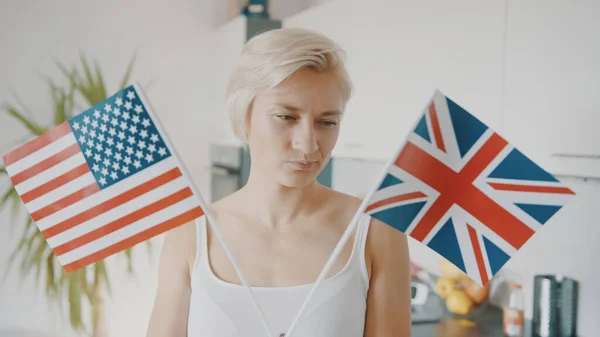 Mujer rubia confundida con banderas de Estados Unidos y Reino Unido en su apartamento. Viajes por trabajo o estudio —  Fotos de Stock