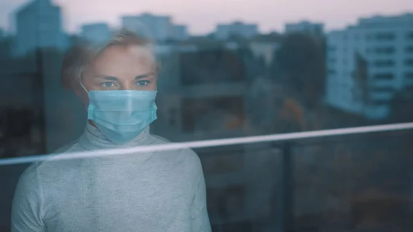 Mujer joven y ansiosa con mascarilla mirando desesperadamente por la ventana. Problema de salud mental durante la cuarentena — Foto de Stock