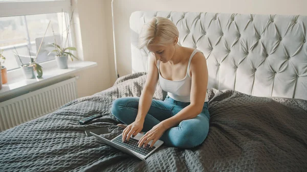 Mujer caucásica joven usando el ordenador portátil en su dormitorio. Freelancer trabajando desde la comodidad de su hogar. — Foto de Stock