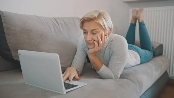 Joven mujer dura charlando con amigos en las redes sociales utilizando el ordenador portátil en su sofá. — Foto de Stock