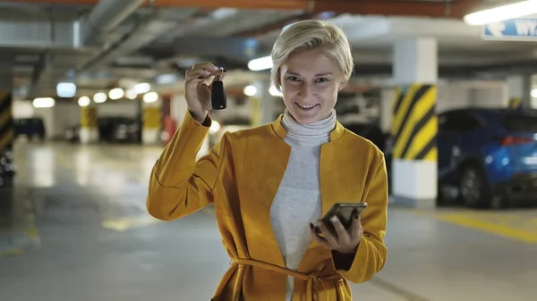 Jovem loira elegante mulher segurando chaves do carro e smartphone na garagem de estacionamento subterrânea — Fotografia de Stock