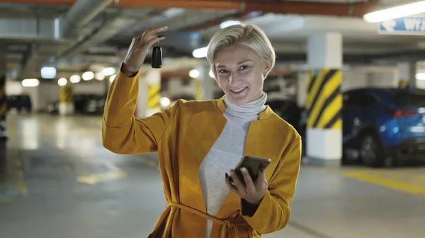 Jovem loira elegante mulher segurando chaves do carro e smartphone na garagem de estacionamento subterrânea — Fotografia de Stock