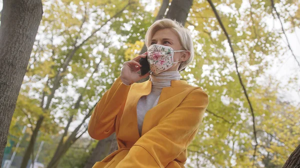 Junge modische Frau mit Stoffgesichtsmaske telefoniert an einem Herbsttag im Park unter dem Baum — Stockfoto