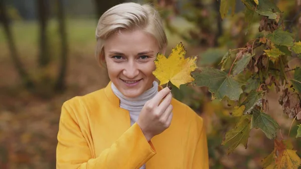 Portraitaufnahme einer jungen kaukasischen blonden Frau mit blauen Augen, die ihr Gesicht mit gelbem Herbstblatt verdeckt — Stockfoto