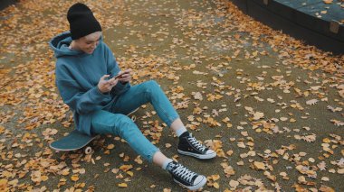 Genç hippi kadın sonbaharda parktaki klavyede oturuyor ve telefonuyla oyun oynuyor.