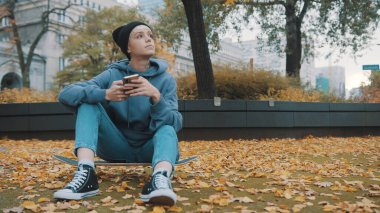 Sonbahar yapraklarıyla kaplı parkta kaykay yaparken telefonu kullanan beyaz tenli genç bir kadın.