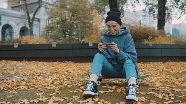 Jovem hipster mulher sentada no tabuleiro de dispersão e jogando jogos em seu telefone no parque no outono — Fotografia de Stock