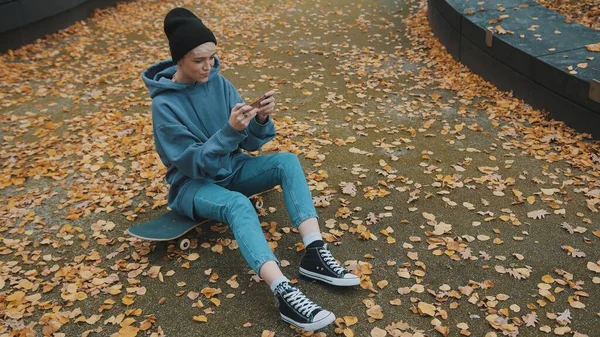 Joven mujer hipster sentada en el scateboard y jugando juegos en su teléfono en el parque en otoño — Foto de Stock