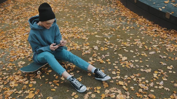 Joven mujer hipster sentada en el monopatín y jugando juegos en su teléfono en el parque en otoño. Disparo de ángulo alto — Foto de Stock