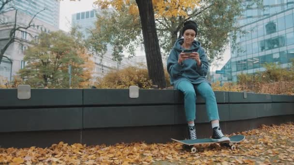 Осенью в городском парке сидит молодая хипстерша со скейтбордом и телефоном. Девушка играет в игру на смартфоне — стоковое видео