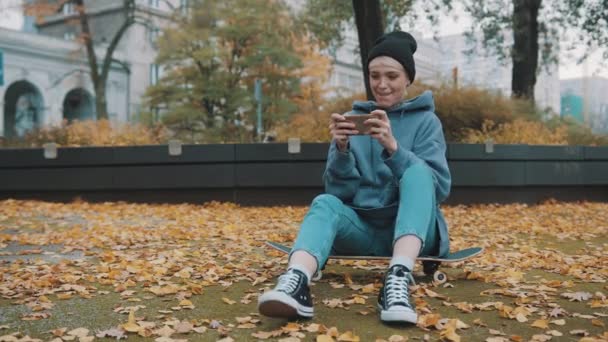 Νεαρή γυναίκα hipster κάθεται στο skateboard και παίζει παιχνίδια στο τηλέφωνό της στο πάρκο στο outside — Αρχείο Βίντεο