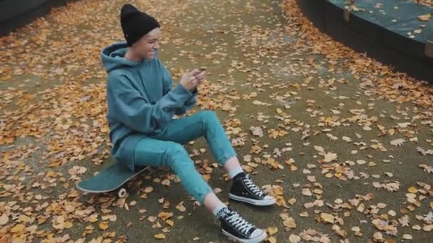 Jonge Kaukasische vrouw met zwarte winter hoed spelen van games n smartphone terwijl zitten op skateboard omgeven door gele herfst bladeren — Stockvideo