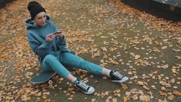 검은 색 겨울 모자를 쓰고 노란 단풍 잎으로 둘러싸인 스케이트보드 위에 앉아 스마트폰으로 게임을 하고 있는 젊은 코카서스 여성 — 비디오