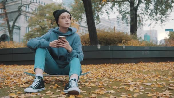 Mladý kavkazský hipster žena pomocí telefonu v parku pokryté podzimními listy, zatímco sedí na skateboardu — Stock video