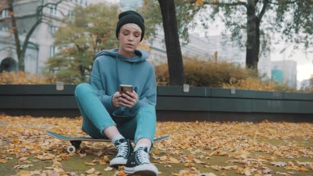 Sonbahar yapraklarıyla kaplı bir şekilde kaykayın üzerinde otururken parkta selfie çekmek için telefon kullanan beyaz tenli genç kadın. — Stok video