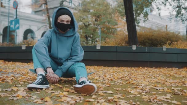 Ung hispter kvinna med ansiktsmask och luvtröja sitter på skateboard i stadsparken på hösten — Stockvideo