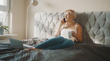 Genç sarışın kadın yatak odasında çay ya da kahve içerken telefonla konuşuyor.. 