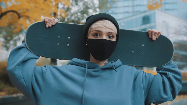 Retrato de una joven patinadora caucásica con máscara protectora que sostiene el monopatín detrás de su cabeza en el parque en otoño — Foto de Stock
