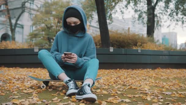 Junge Kaukasierin mit Kapuzenpulli und Gesichtsmaske sitzt am Herbsttag auf dem Skateboard und nutzt Smartphone — Stockvideo